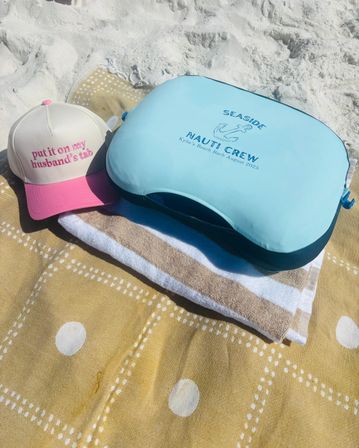 Sunny white-sand beach scene: a pink-and-cream trucker hat embroidered 'put it on my husband's tab' sits beside a light-blue cooler bag with anchor graphic and 'Nauti Crew' text on striped beach towels.