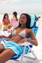 Young woman sunbathing in a striped bikini and blue shorts on a striped beach chair at a sunny sandy beach, friends chatting nearby by the shoreline.