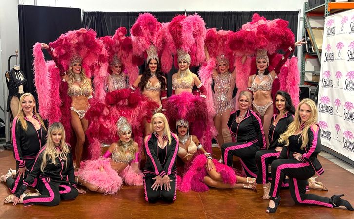 Backstage group of Las Vegas‑style showgirls and crew posing in bright pink feather headdresses, rhinestone bikinis and matching black-and-pink tracksuits, holding large feather fans.