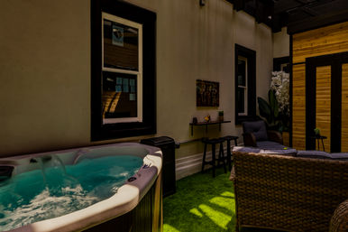 Cozy outdoor patio with bubbling hot tub, wicker lounge seating and cushions, bar stools, potted plants and a wooden privacy screen on green artificial turf.