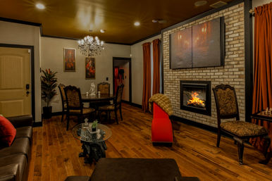 Cozy warm-toned living room with brick fireplace and roaring fire, oversized wall art, crystal chandelier, hardwood floors, ornate dining chairs and rust-colored curtains