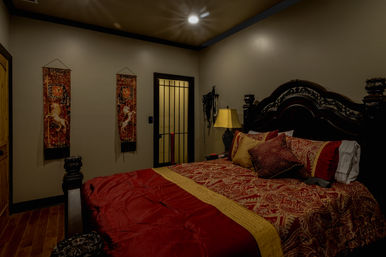 Luxurious master bedroom interior with ornate dark wood headboard, red-and-gold paisley bedding, tapestry wall hangings, hardwood floor and warm bedside lamp