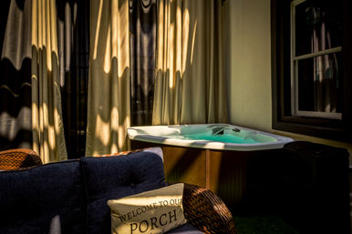 Sunlit outdoor porch with a bubbling hot tub, beige curtains casting wavy shadows, wicker chair and navy sofa with a 'Welcome to our porch' pillow — a relaxing patio scene.