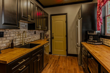 Cozy galley kitchen with matte black cabinets, butcher block countertops, brick backsplash, brass hardware and a mustard-yellow paneled door, stainless appliances, coffee maker, warm hardwood floors and a red patterned window valance.