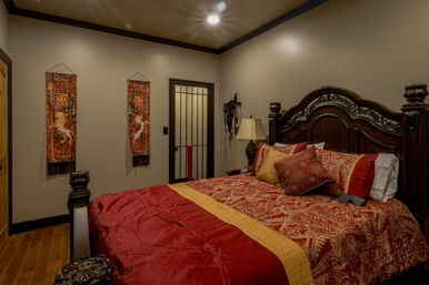 Traditional luxury bedroom with ornate dark wood headboard, red and gold paisley bedding and throw pillows, carved nightstand with lamp, decorative horse tapestries on beige walls, hardwood floors and recessed lighting.