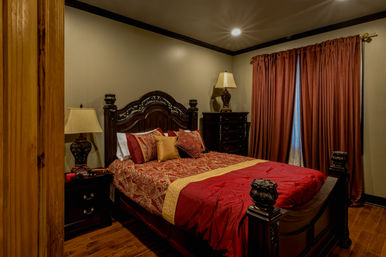 Cozy luxury master bedroom with ornate dark wood bed and carved headboard, red and gold patterned bedding, matching nightstands and decorative lamps, hardwood floors and rust-colored floor-length curtains under warm recessed lighting.