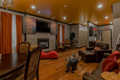 Cozy luxury lounge interior with brick fireplace and wall-mounted TV, orange velvet curtains, dark leather seating, hardwood floors and ornate chandelier.