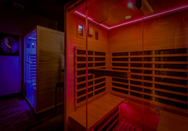 Glowing pink-lit infrared wooden sauna with glass door and bench seating, adjacent to a blue-lit steam shower in a modern spa room.