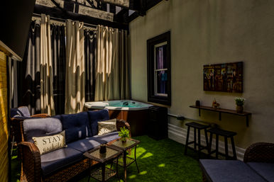 Cozy covered patio with wicker sofa and blue cushions, small glass-top tables, privacy curtains, hot tub, bar-style counter with stools, wall art and artificial grass flooring