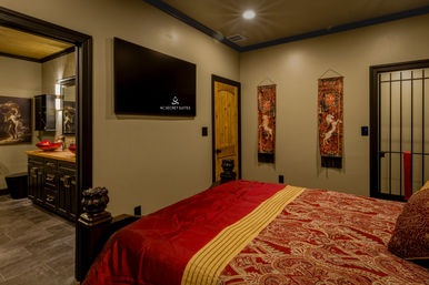 Cozy boutique bedroom suite with red paisley bedding, wall-mounted flat-screen TV, wooden door, decorative unicorn tapestries, and a dark vanity with red vessel sinks visible through the doorway under warm ambient lighting