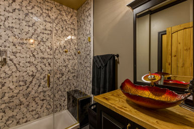 Residential bathroom interior with marble hexagon mosaic shower and glass door, brass fixtures, dark towel, wooden vanity topped with a red glass vessel sink and mirror under warm lighting.