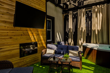 Sunlit cozy patio with wood-paneled wall and mounted TV, wicker sofa with blue cushions and 'stay awhile' pillow, small wicker tables, privacy curtains and a hot tub on green artificial grass