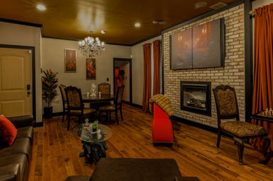 Cozy living room with crystal chandelier, round dining table and ornate chairs, brick fireplace, hardwood floors and rust-red curtains.