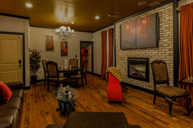 Cozy living room with crystal chandelier, round dining table and ornate chairs, brick fireplace, hardwood floors and rust-red curtains.