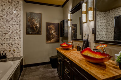 Stylish luxury bathroom with eye-catching red glass vessel sinks on a wooden vanity, black cabinetry, marble mosaic tile wall, illuminated mirror and framed classical paintings.