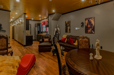 Cozy upscale living room and open kitchen interior with hardwood floors, brown leather sofa and red pillows, ornate dining chairs, red chaise lounge, decorative suit of armor, framed artwork and warm recessed lighting.