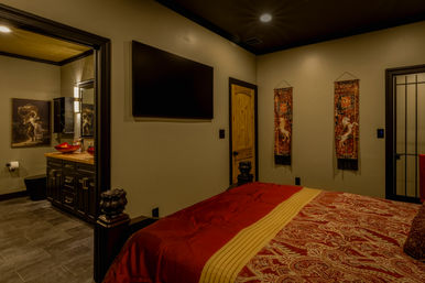 Warm modern-rustic master bedroom with red paisley bedding, wall-mounted flat-screen TV, wooden door and decorative tapestries, plus an adjoining en-suite bathroom featuring red vessel sinks, dark wood vanity and slate tile floor.