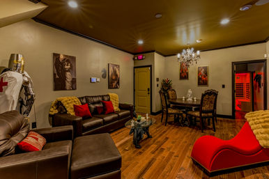 Residential basement lounge with dark leather sofas, red chaise, suit of armor, crystal chandelier, dining table, wall art and hardwood floors with a red-lit sauna visible through an open door.