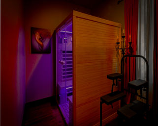 Cozy spa treatment room with a wooden infrared sauna cabin glowing purple, red curtains, decorative candelabra and black therapy stools for relaxation.