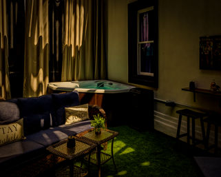 Cozy sunlit indoor patio with a bubbling hot tub, wicker sofa with navy cushions, glass-top wicker tables on green turf and drawn curtains.