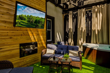Cozy covered outdoor patio with wood-paneled wall and wall-mounted TV showing a green landscape, wicker loveseat with blue cushions and decorative pillows, small wicker tables, privacy curtains, and a hot tub on artificial turf.