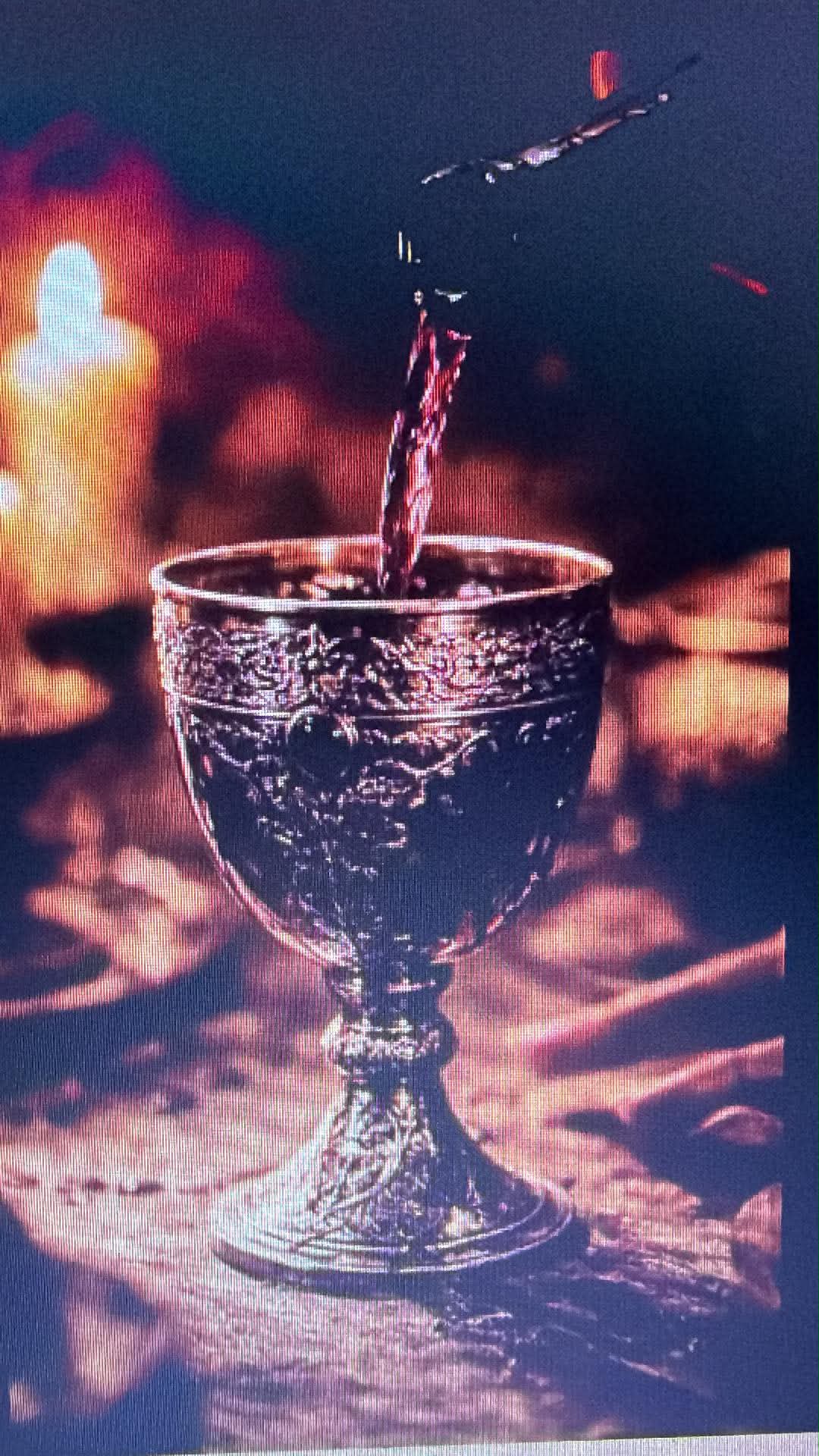 Candlelit close-up of an ornate decorative goblet being filled with red wine, warm golden bokeh and textured table