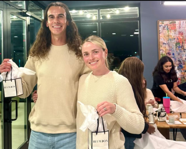 Two smiling friends in matching cream sweaters holding small white gift bags at a cozy indoor party in a modern apartment, with people chatting around a table in the background.