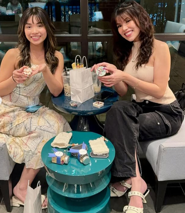 Two friends smiling at a cozy cafe table with bubble tea, gift bags and decorated desserts on a teal side table by a nighttime city window