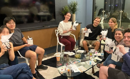 Group of friends in a modern apartment living room having a casual gift exchange around a glass coffee table, each holding small white-tissue wrapped gift boxes, with potted plants and balcony doors in the background.