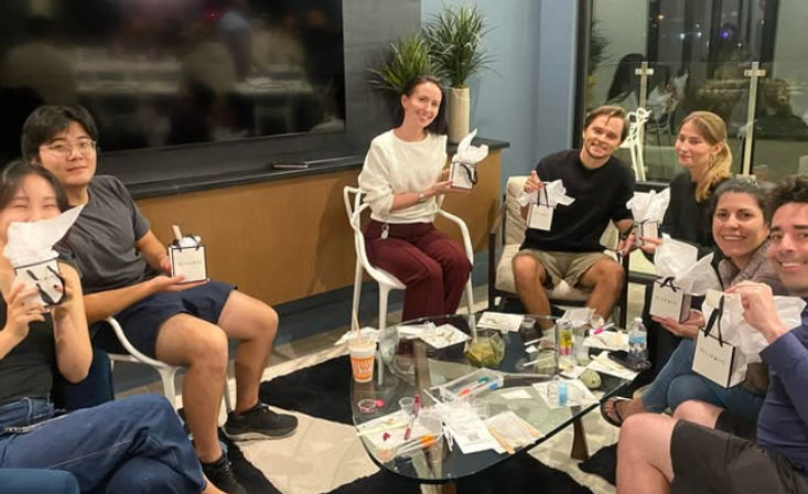Group of friends in a modern apartment living room having a casual gift exchange around a glass coffee table, each holding small white-tissue wrapped gift boxes, with potted plants and balcony doors in the background.