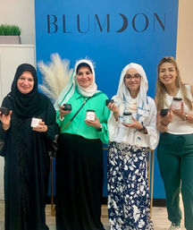 Four smiling women, two wearing headscarves, pose indoors against a bright blue wall holding small jar candles and lids — friendly group photo in a boutique wellness/gift shop.