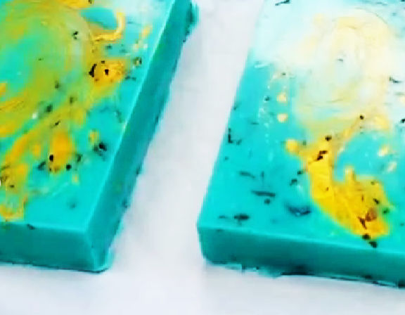 Two handcrafted turquoise soap bars with bright yellow-gold marbled swirls and tiny black speckles displayed on a white surface