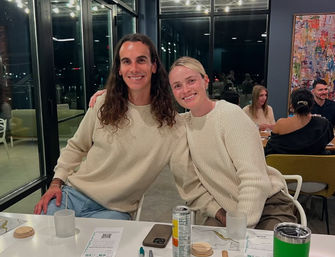 Two smiling friends in matching cream sweaters sit close together at a modern glass-walled restaurant at night, one with long dark hair and one with short blonde hair, with menus, drinks and a phone on the table and city lights visible through large windows.