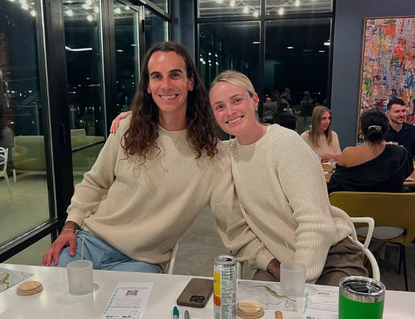 Two smiling friends in matching cream sweaters sit close together at a modern glass-walled restaurant at night, one with long dark hair and one with short blonde hair, with menus, drinks and a phone on the table and city lights visible through large windows.