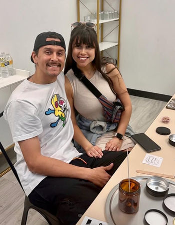 Smiling couple seated at a bright craft studio table, making a candle with an amber jar and wick while phone and candle-making supplies sit on the work surface.