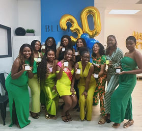 Group of women in green outfits posing indoors for a 30th birthday celebration with large gold "30" balloons and blue balloons, smiling and holding candles or small gift jars.