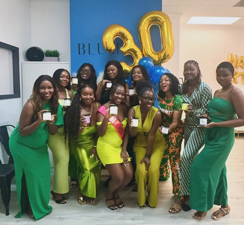 Group of women in green outfits posing indoors for a 30th birthday celebration with large gold "30" balloons and blue balloons, smiling and holding candles or small gift jars.
