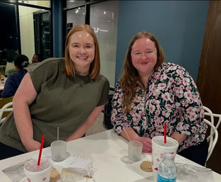 Two smiling friends seated at a white table in a modern indoor dining area with drinks (red straws), a water bottle and wrapped cookies on the table.