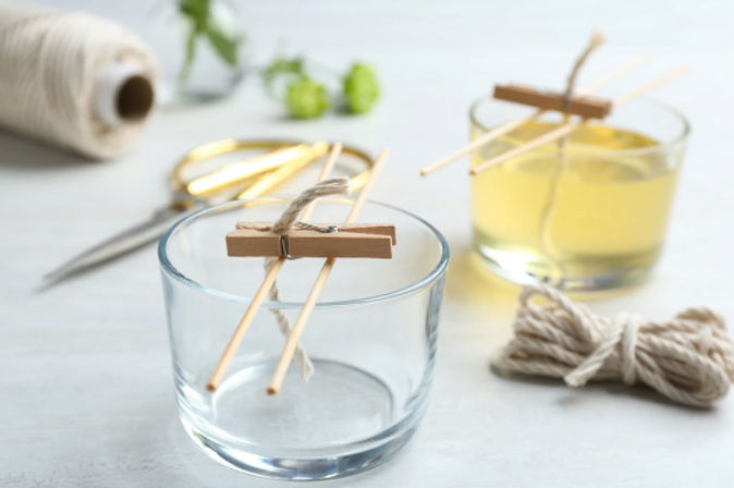 DIY candle-making scene: clear glass votive jars with cotton wicks held by mini clothespins and wooden skewers, jar of yellow melted wax, twine and scissors on a bright craft table.