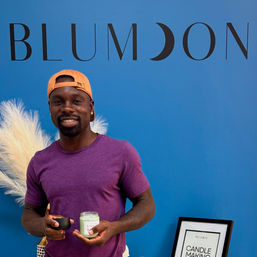 Smiling man in a purple shirt and orange cap holding a small jar candle and lid in front of a bright blue wall with pampas grass and a framed candle-making sign.