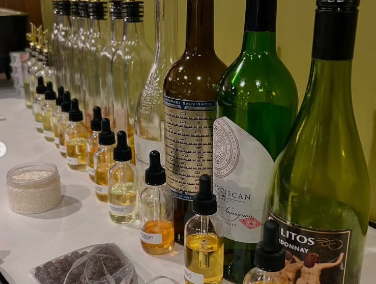 Lineup of wine bottles and small labeled dropper bottles filled with golden and clear liquids, plus a jar of powder and a bag of chocolate chips on a white table — a wine-tasting or cocktail-flavoring setup.