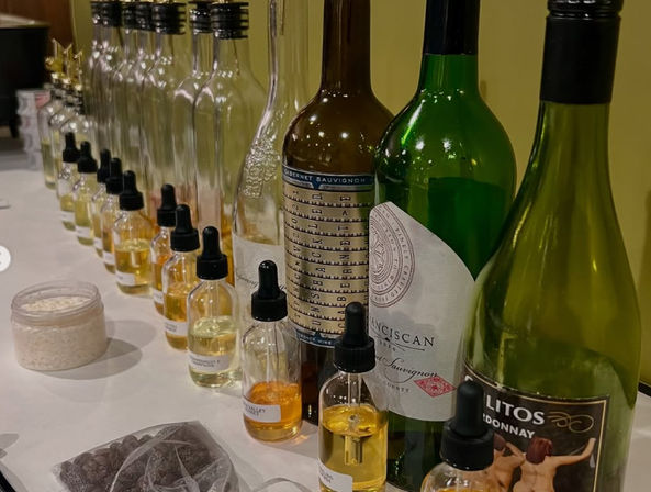 Lineup of wine bottles and small labeled dropper bottles filled with golden and clear liquids, plus a jar of powder and a bag of chocolate chips on a white table — a wine-tasting or cocktail-flavoring setup.