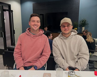 Two smiling men in hoodies sitting at a table in a modern coworking lounge, casual meeting with drinks, a phone, a can, wall-mounted TV and potted plants in the background.