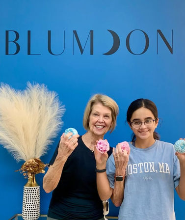Two smiling people holding colorful bath bombs in a bright blue boutique with pampas grass; one wears a Boston, MA T-shirt.