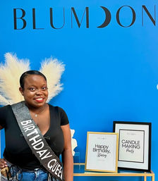 Smiling woman wearing a 'Birthday Queen' sash in front of a bright blue backdrop with decorative white feather plumes and framed event signs for a birthday and candle-making party.