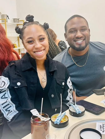 Two smiling people at a bright indoor candle-making workshop, mason jar candle and small tins with wicks and clothespin holders on the table.