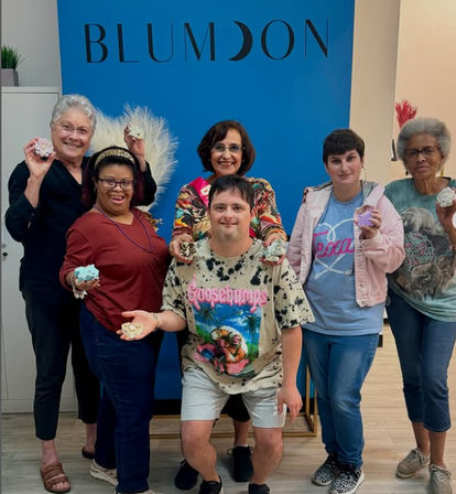 Smiling diverse group of six adults posing indoors at a community craft workshop, each holding colorful handmade bath bombs or soaps against a bright blue backdrop.