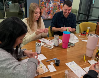 Smiling group of people around a wooden table in an office-style meeting room doing an after-work craft night, folding paper and writing with pens amid canned drinks, colorful tumblers and small craft supplies.