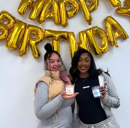Two smiling friends holding small candle jars under shiny gold "HAPPY BIRTHDAY" balloon letters at an indoor celebration.
