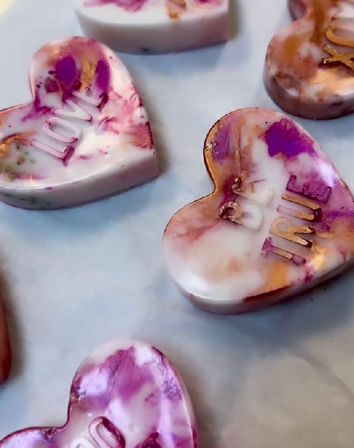 Close-up of handmade marbled heart-shaped soaps with pink, purple and gold swirls and embossed "LOVE" lettering on parchment paper.
